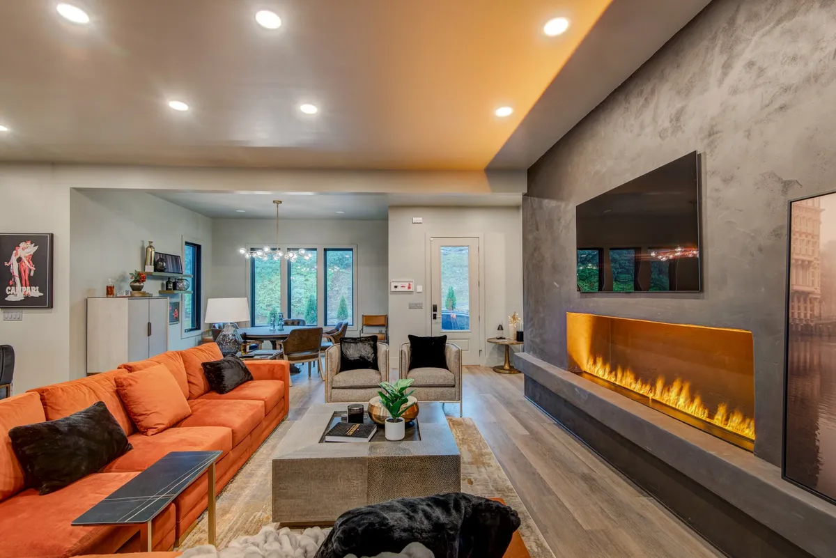 An open-concept living and dining area featuring an orange sectional, modern fireplace, expansive windows with mountain views, and a stunning chandelier above the dining table. The scene exudes warmth and contemporary style, perfect for both relaxation and entertaining guests.