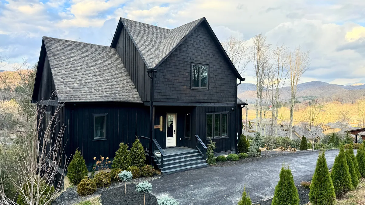 A modern black exterior of the house surrounded by a landscaped garden, with a pathway leading to the entrance. The scene feels welcoming and contemporary, suggesting a connection to the outdoors and the potential for gatherings in all seasons.