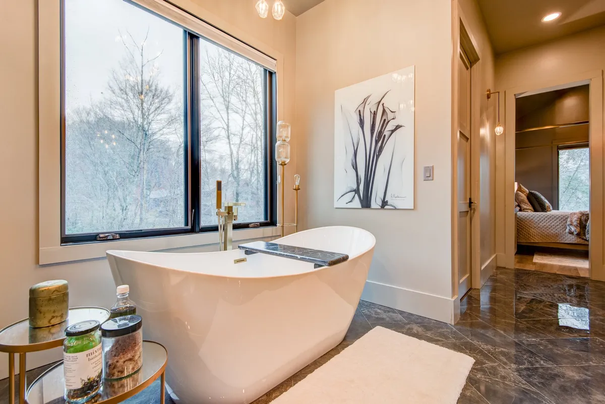 A luxurious bathroom with a freestanding tub positioned near large windows that flood the space with natural light. Marble flooring and abstract wall art add to the spa-like ambiance, creating a serene and indulgent retreat.