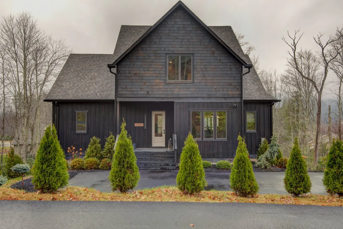The house's dark wooden facade with a landscaped garden and front steps, exuding calmness and a sense of connection to nature. The porch area hints at enjoying the tranquility and beauty of the changing seasons, from vibrant fall foliage to summer evenings outside.