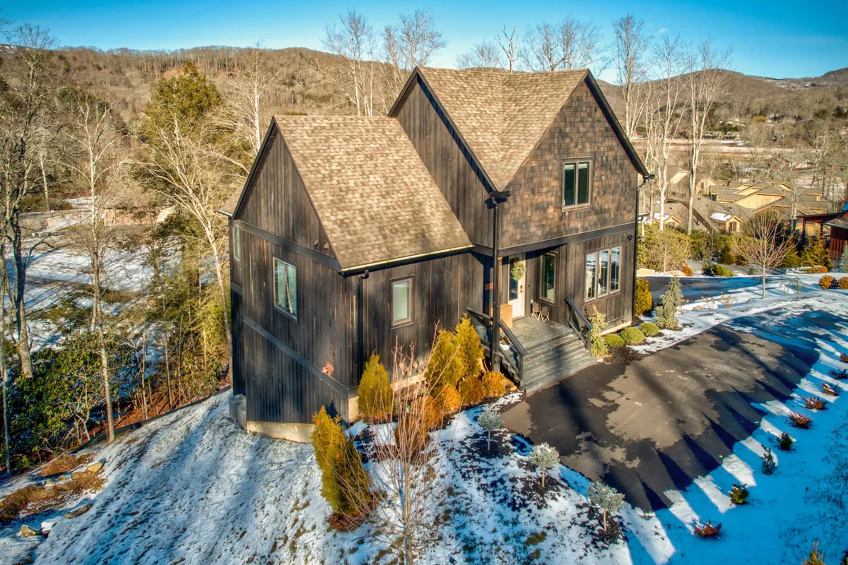 The house is surrounded by a manicured garden and mature trees, blending seamlessly into the natural landscape. The scene captures the welcoming exterior, offering glimpses of outdoor living space and the wooded environment that waits to be explored.