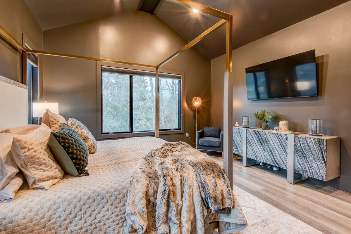 A spacious bedroom featuring a canopy bed, large windows letting in natural light, a wall-mounted TV for private entertainment, and a decorative console table. The room feels both comfortable and inviting, emphasizing personal relaxation and modern amenities.