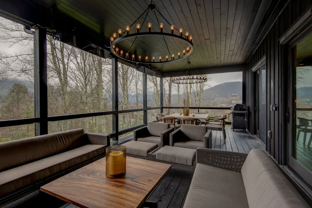 An inviting outdoor deck with a stylish dining table and comfortable seating, surrounded by trees. Elegant chandeliers add ambience for evening gatherings, while the open setting offers glimpses of the surrounding landscape.