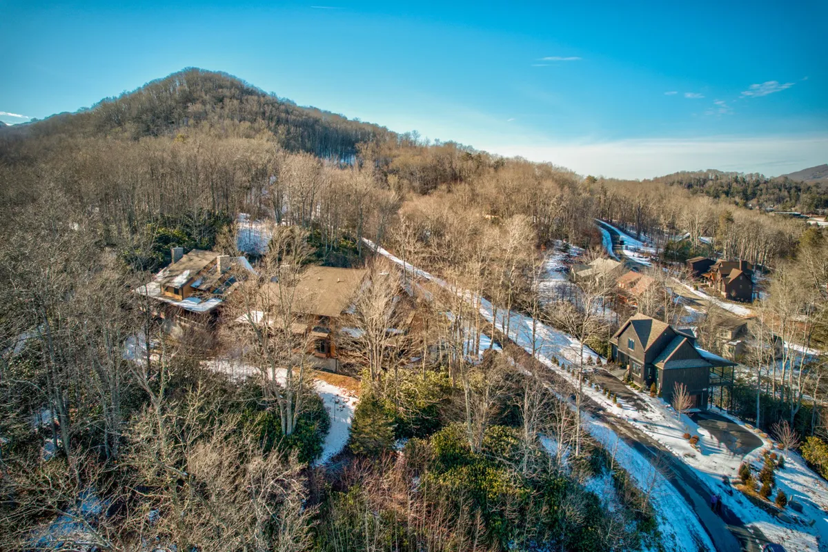A sprawling house is nestled among mature trees with a winding road leading through a scenic landscape and dramatic mountain backdrop. The greenery and expansive views evoke a sense of adventure and connection to nature, highlighting the property's proximity to outdoor activities and Banner Elk's natural beauty.