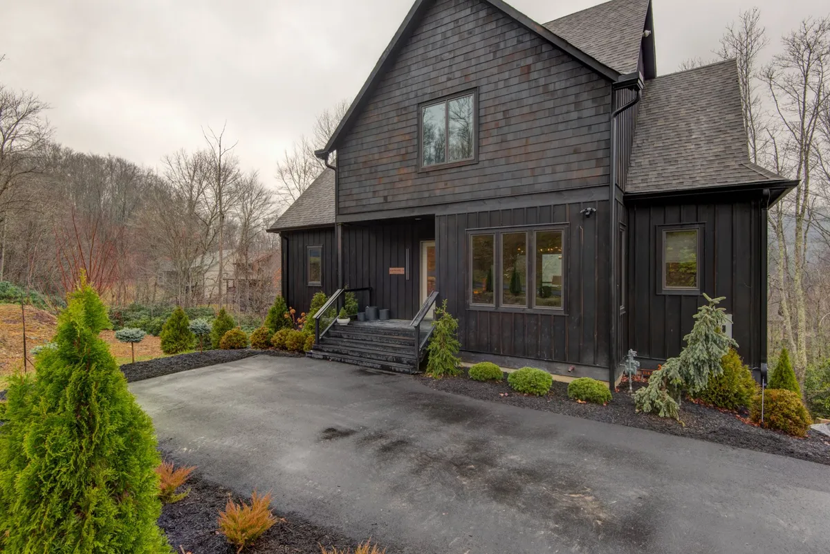 This exterior image showcases the striking dark wooden facade of the house, welcoming front steps, large windows, and lush greenery surrounding the property. The setting is contemporary but cozy, blending the home seamlessly into its natural environment while offering a comforting sense of privacy.