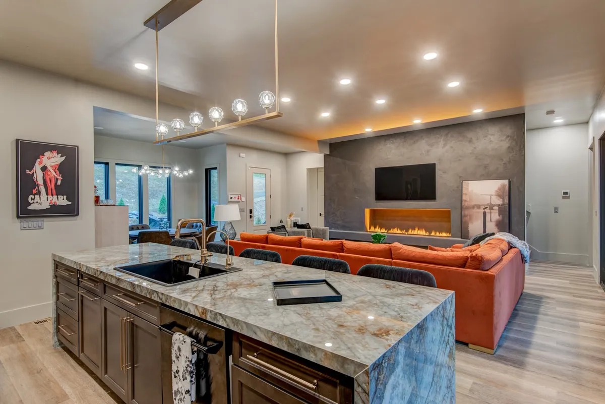 A wide-angle view blending the living area, modern kitchen island, vibrant artwork, and a central fireplace. The orange sectional sofa anchors the room, while the kitchen’s proximity highlights easy circulation and socializing.