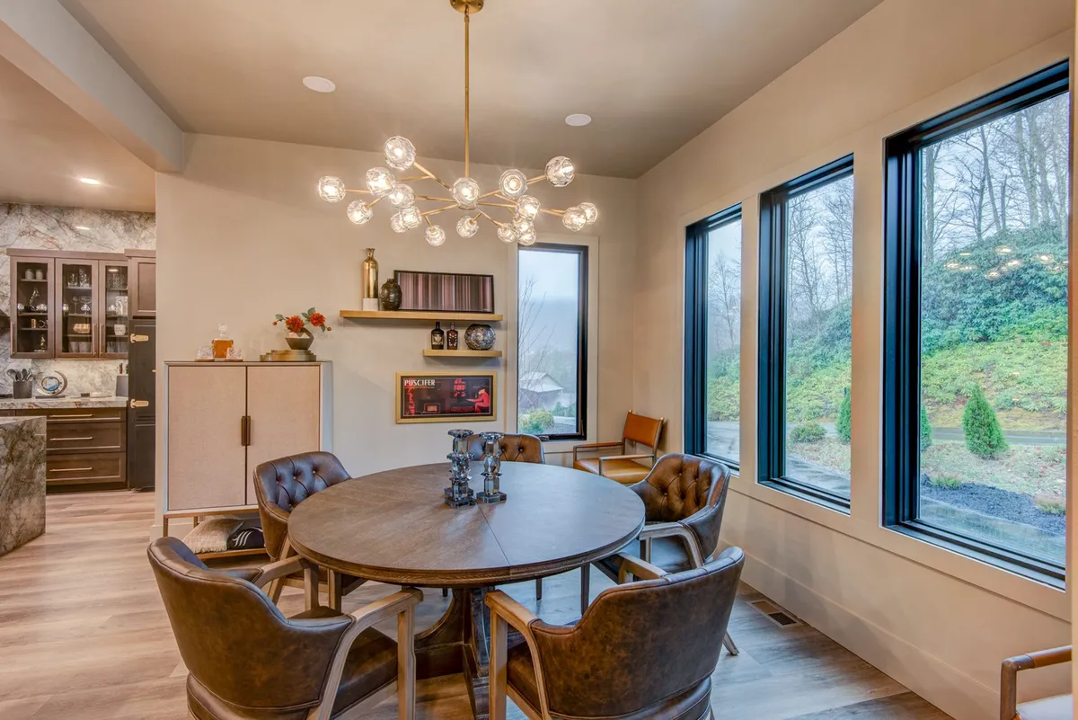 A chic, inviting dining area with a round table and chandelier, set beside large windows that reveal scenic views. The adjacent kitchen cabinetry is visible, tying the dining space back to the kitchen's atmosphere of culinary activity. The mood is cozy yet refined, perfect for both intimate family meals and celebratory feasts.