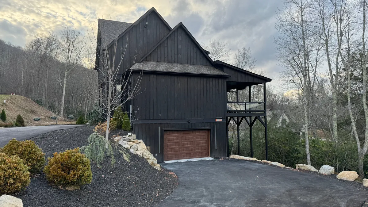 An exterior shot featuring a dark wooden house, an elevated porch, a landscaped pathway, and surrounding trees. The setting is tranquil, and the porch offers a cozy vantage point to enjoy the outdoors, particularly by a firepit.