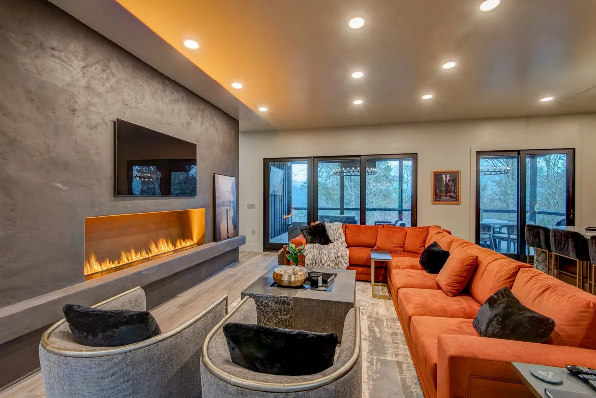 A spacious living room with a large orange sectional sofa, a sleek modern fireplace, a flat-screen TV, and expansive viewing windows that offer glimpses of mountain vistas. The high ceilings and open layout create an airy, inviting atmosphere ideal for gatherings.