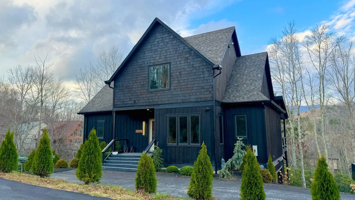 The image features the striking black wooden facade of a modern single-family home set against the lush, landscaped front yard. Elegant steps lead to the inviting entrance, and the serene surroundings hint at the tranquility and luxury inside. The setting is perfect for gathering outdoors or enjoying peaceful mountain evenings.