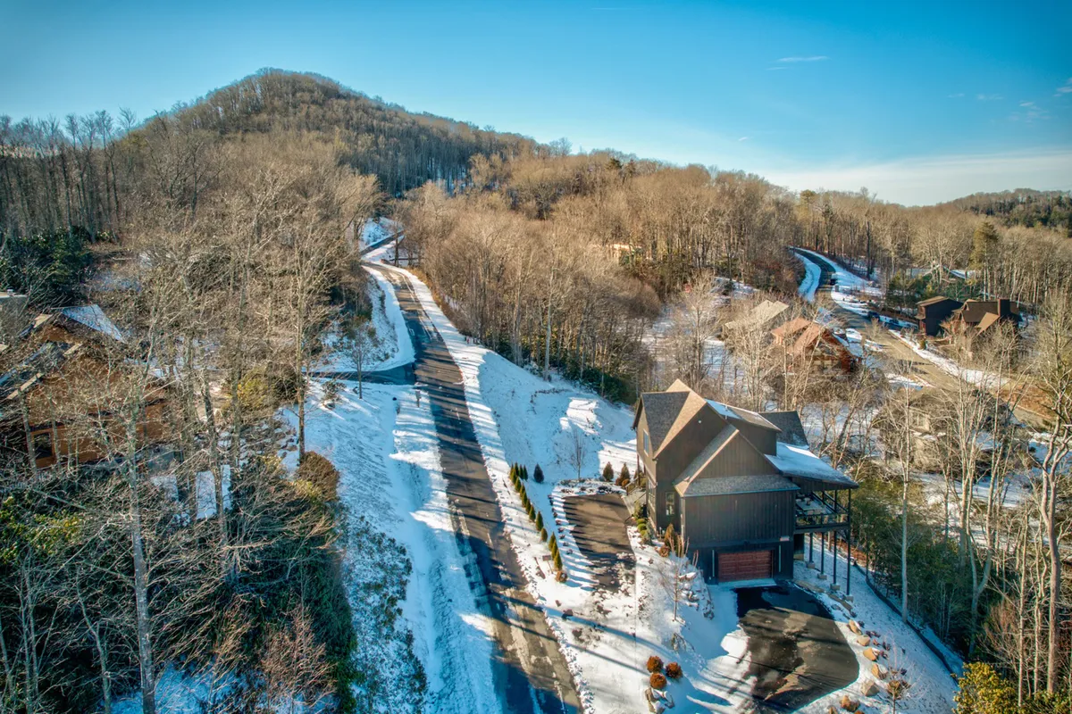 Exterior view of the house, surrounded by lush trees and a winding road, with expansive mountain vistas in the background. The scene conveys a sense of privacy, tranquility, and accessibility to nearby outdoor adventures.