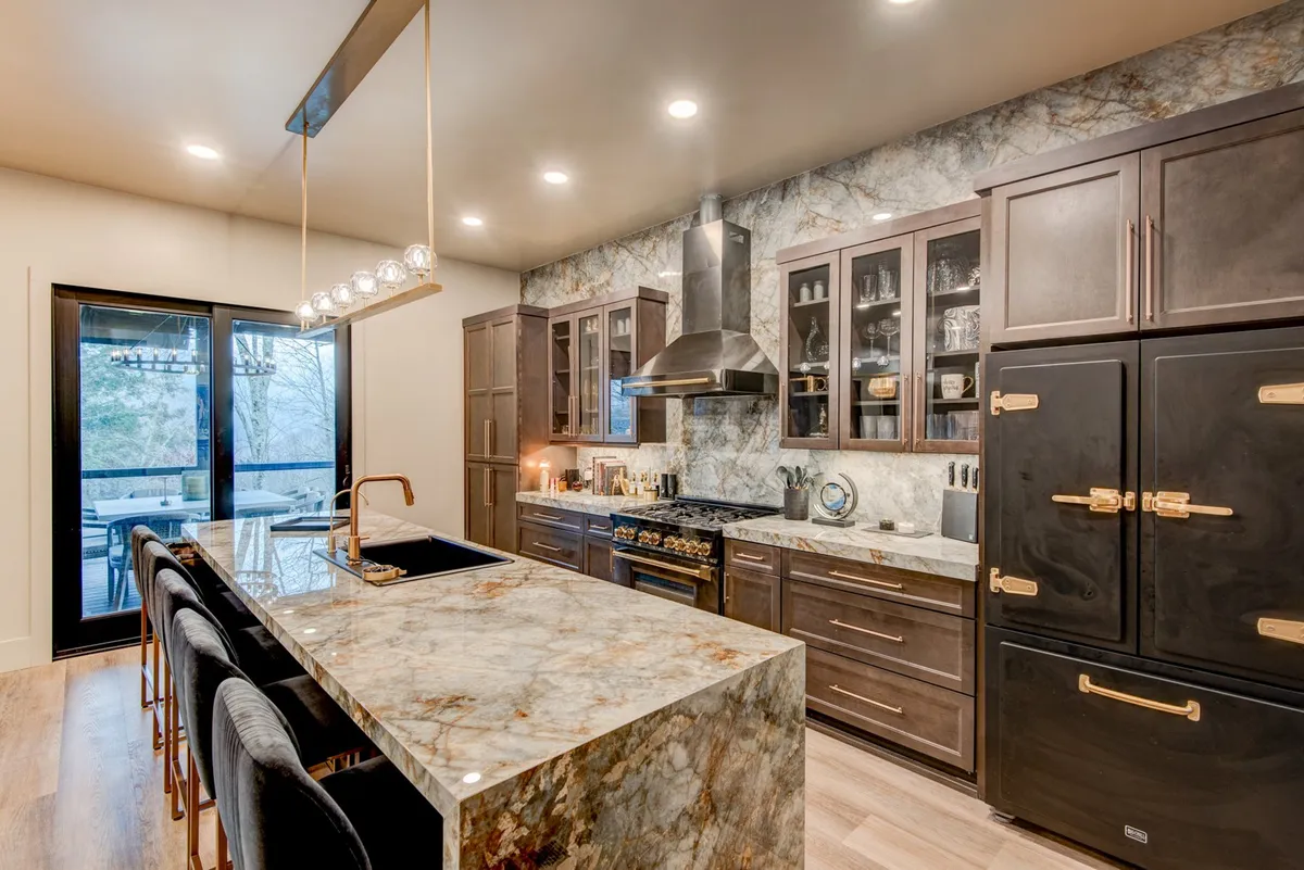 A spacious, modern kitchen featuring a large island topped with sleek porcelain, surrounded by bar seating. Stainless steel Zline appliances, glossy cabinetry, and decorative lighting fixtures give the space an upscale, sophisticated feel. The open layout hints at both functionality for serious cooking and an inviting atmosphere for gatherings.